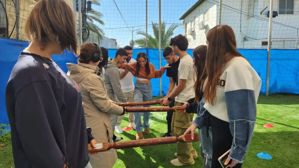 תלמידות ותלמידים מחזיקים מוטות עץ ומלווים חברה לקבוצה במשימת אמון על מגרש דשא