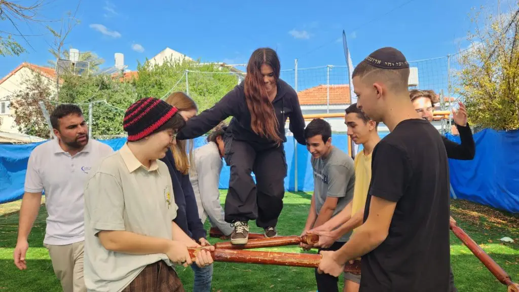 תלמידים בפעילות גיבוש ODT — תחנת שיווי משקל: נערה הולכת על מוטות עץ שהקבוצה מחזיקה יחד, בהנחיית דניאל חסיד